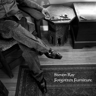 A square lack and white photo. A figure sits in a chair in a wooden chair in the upper left corner. Head and shoulders are out of the frame. They are dressed up in nice shirt and pants with fancy black shoes. Their legs are crossed. Their left hand is reaching toward a small electrical device on an octagonal wooden moroccan table. Their index finger is about to press on a button on the device. There is a shadow of their hand with the finger pointing down on the wall behind them. To the right of the table is another piece of wooden furniture with another device on it. The wooden floor is visible and a rug covers the bottom third of the floor. Near the bottom right in a fancy font is the white text Steven Ray Forgotten Furniture.